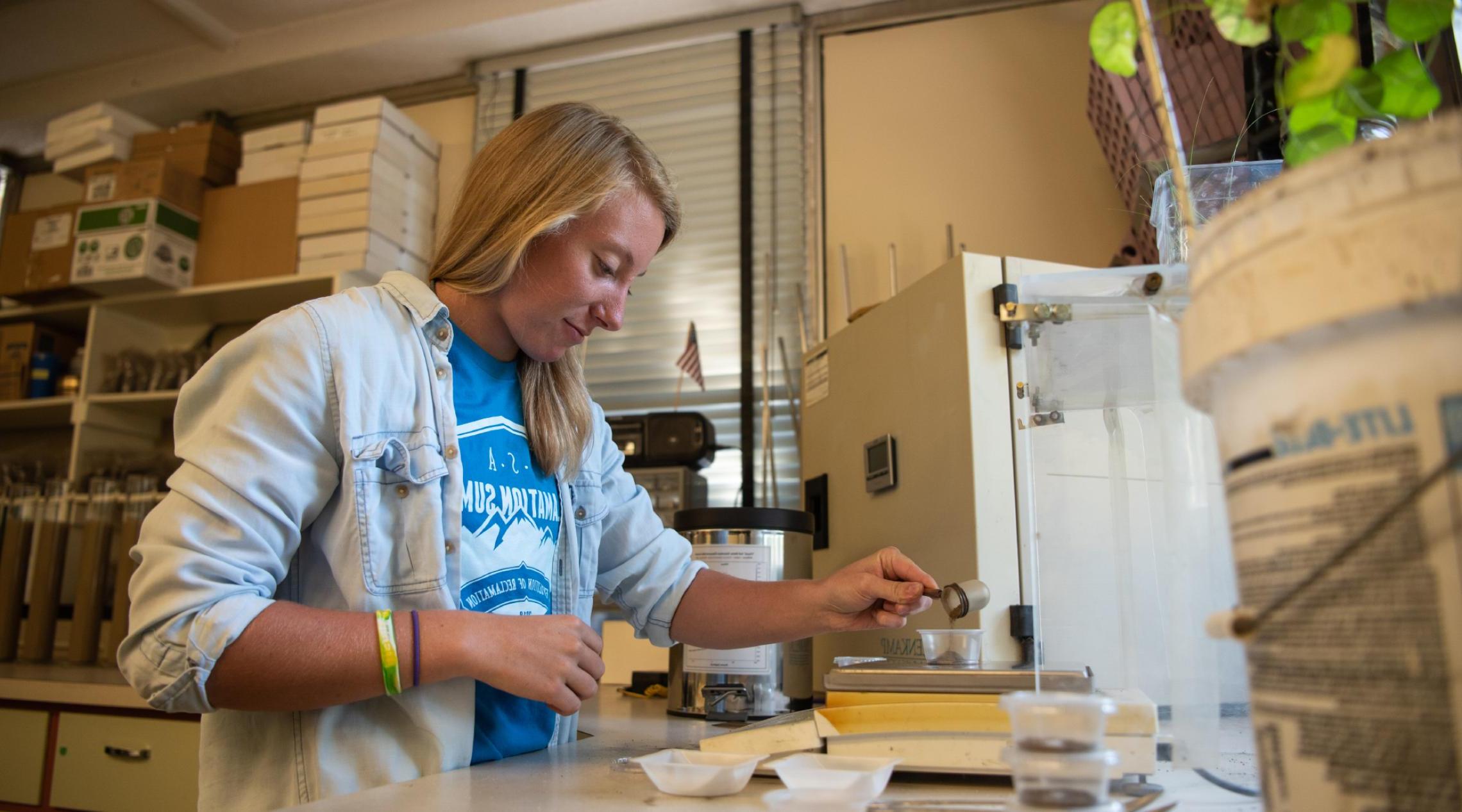 A young woman in a lab pours a small amount of soil into a clear plastic cup.